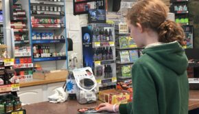 Shows middle school aged child at convenience store counter purchasing a candy bar. On the counter are CBD vape liquids and lighters. Behind the counter is a large display of tobacco products.